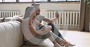 Happy young couple holding phone taking selfie sit on floor