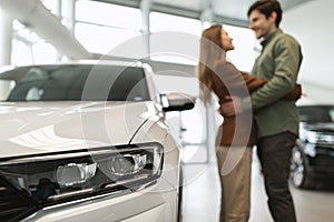 Happy young couple hugging at auto dealership, celebrating purchase of new vehicle, selective focus on car