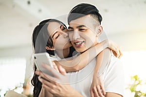 Happy young couple kissing and  looking at mobile phone in living room at home