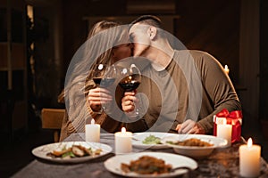 Happy young couple in love kissing, having romantic dinner celebrating Valentines day, drinking red wine at home or in