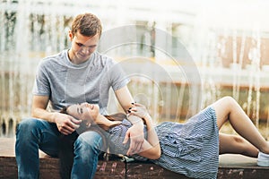 Happy young couple in love teenagers friends dressed in casual style sitting together near fountain on city street