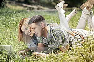 Happy young couple lying on the grass, looking at laptop and smiling. Relationship and learning concept.