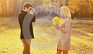 Happy young couple, man photographs woman with film camera and yellow maple leaves in autumn park on warm sunny day