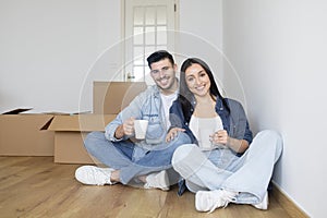 Happy young couple moving home, sitting among cardboard boxes and drinking coffee