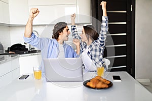 Happy young couple with raised arms of win celebrate looking on laptop in kitchen