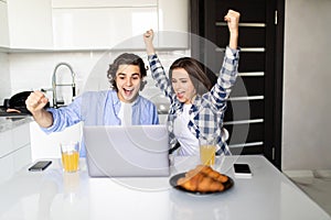 Happy young couple with raised arms of win celebrate looking on laptop in kitchen