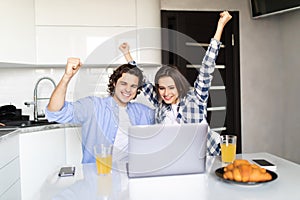 Happy young couple with raised arms of win celebrate looking on laptop in kitchen