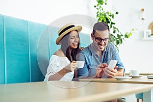 Happy young couple sitting at the cafe table drinking coffee and looking at mobile phone