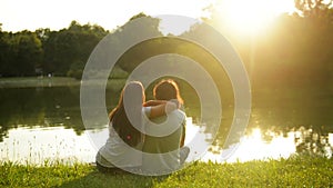 Happy young couple sitting on the green grass near the lake during sunset. Back view of woman and man in love outdoors.