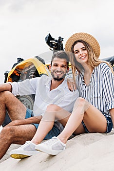 happy young couple sitting near ATV on sandy dune and looking