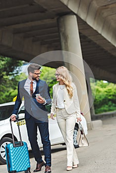 happy young couple smiling each other while going with suitcase near taxi