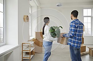 Happy young couple standing back with cardboard boxes and a plant in a new apartment on moving day.