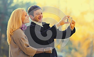 Happy young couple taking selfie with mobile phone in autumn park on warm sunny day
