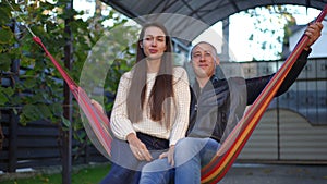 Happy young couple with toothy smile sitting on hanging hammock enjoying weekend outdoors. Positive Caucasian man and