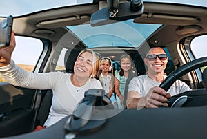 Happy young couple with two daughters inside car during auto trop. They are smiling, laughing and taking selfies using smartphone