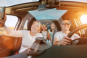 Happy young couple with two daughters inside car during auto trop. They are smiling, laughing and taking selfies using smartphone photo