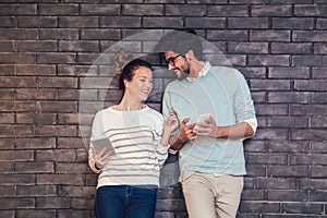 Happy young couple using a tablet and smart phone on a brick wall
