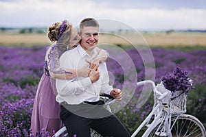 Happy young couple are walking with retro bicycle in lavender field. woman in purple dress and with hairstyle is hugging her man
