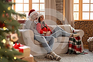 Happy young couple wearing Santa hats on sofa in room decorated for Christmas