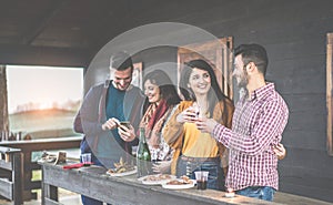 Happy young couples toasting red wine and eating barbecue in backyard wooden house - Cheerful people having fun at sunset in