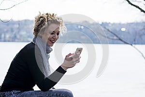 Happy young curly blond woman sitting by the river and typing on