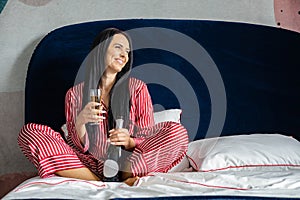 Happy young domestic woman in red pajamas drinking champagne wine glass sitting in comfortable bed