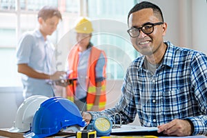 Happy young engineer smiling while working on the desk and checking blueprint have a colleagues as background