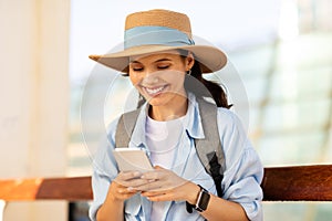 Happy young european lady tourist in hat with backpack, typing on smartphone, use online map, chatting