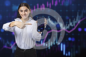 Happy young european woman pointing at cellphone with glowing growing forex chart on blurry background. Trade, finance and stock