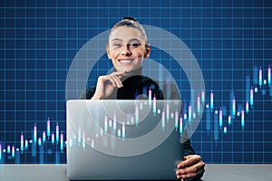 Happy young european woman sitting at desk with laptop and creative growing candlestick forex chart on blue grid background.