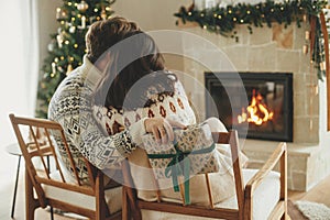 Happy young family in cozy sweaters exchanging stylish christmas gifts on background of fireplace with modern festive mantle and