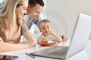 Happy young family with cute kid son eating toasts using laptop in kitchen.
