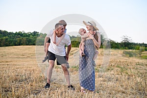 Happy young family. A father, a pregnant mother, and two little sons on their backs. Beveled wheat field on the background. Sunset