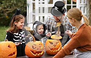 Happy young family in Halloween costumes carving pumpkins together in backyard