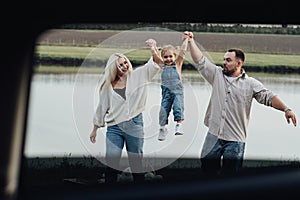 Happy Young Family Having Fun Outdoors, Mom and Dad Taking Up Their Little Daughter by Hands, View Through Window Inside