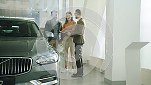 Happy young family husband and wife choosing auto in car dealership talking to salesman