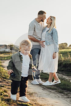 Happy Young Family Outdoors, Father Holding His Pregnant Wife by Belly While Their Toddler Son Playing Around Them