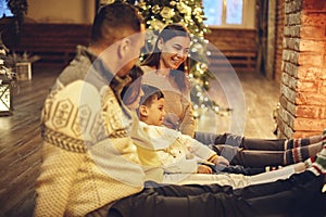 Happy young family parents with two small kids gathering around brick fireplace in cozy and warm country house during Christmas