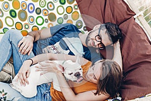 Happy young family portrait, cute stylish woman holding happy bulldog near handsome man in sunglasses while relaxing on a hammock