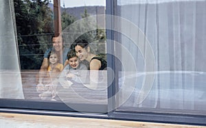 Happy young family is sitting on living room floor. Parents and children hug and have fun at home by glass door