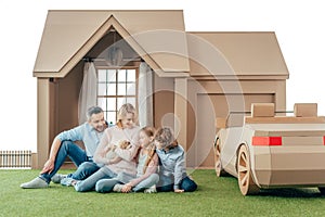 happy young family sitting on yard of cardboard house with their puppy