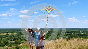 Happy young family of three fly a multi-colored kite, having fun, against background of beautiful summer landscape of