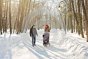 Happy young family walking in the park in winter. The parents carry the baby in a stroller through the snow.