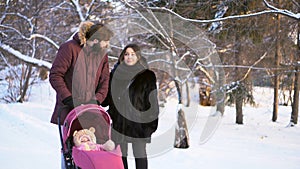 Happy, young family walking in a winter park, mom, dad and baby in pink stroller. Man pointing the finger and parents