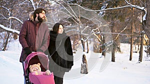 Happy, young family walking in a winter park, mom, dad and baby in pink stroller. Man pointing the finger and parents