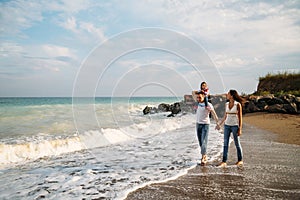 Happy young family in white t-shirts and blue jeans with a small daughter in blue dress walking along the seaside