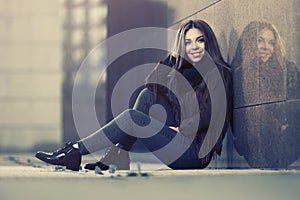 Happy young fashion woman in leather jacket sitting at the wall