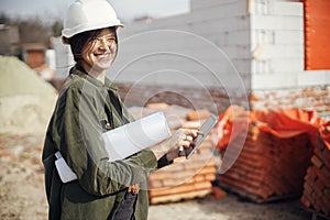 Happy young female architect with tablet checking blueprints against of building new modern house. Stylish happy woman engineer in