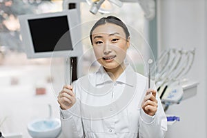 Happy young female asian dentist with tools standing in light modern medical office background.