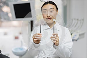 Happy young female asian dentist with tools standing in light modern medical office background.